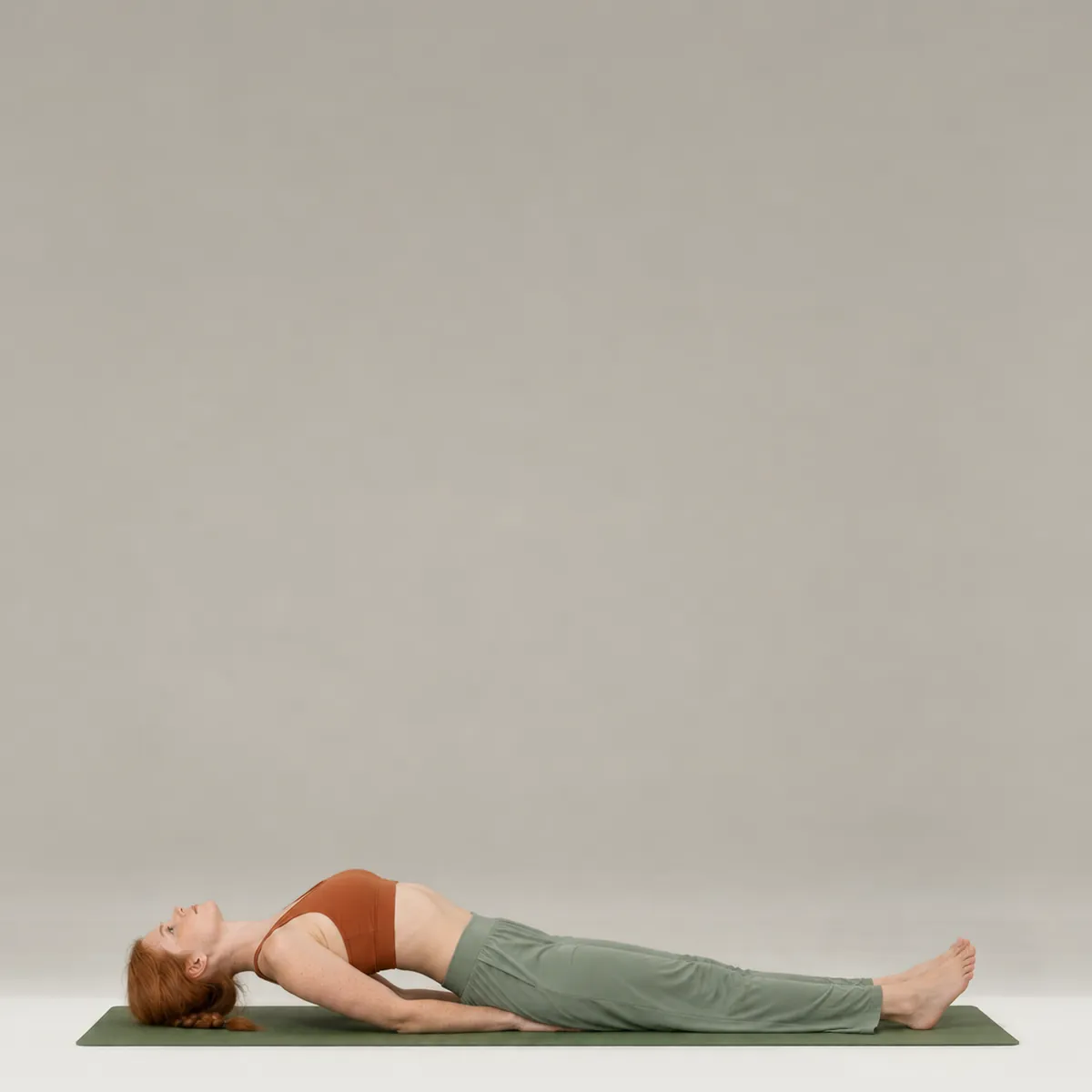 A woman in fish pose on a green yoga mat, lying on her back with her chest lifted