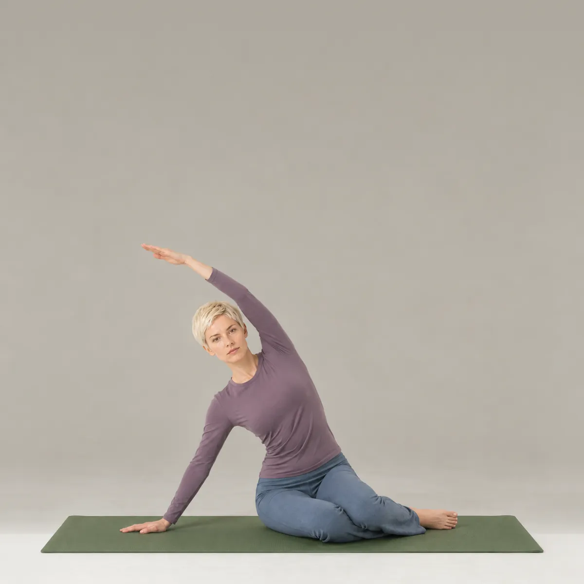 A woman doing the pilates mermaid on a green mat, seated and side bending with one arm overhead