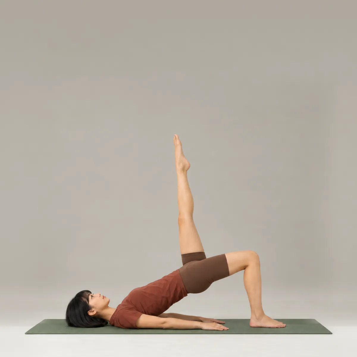 A woman in a single-leg variation of bridge pose on a green yoga mat, hips lifted and one leg reaching up