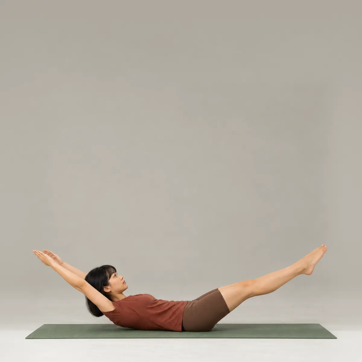 A woman doing the pilates double leg stretch on a green mat, arms reaching back and legs extended forward