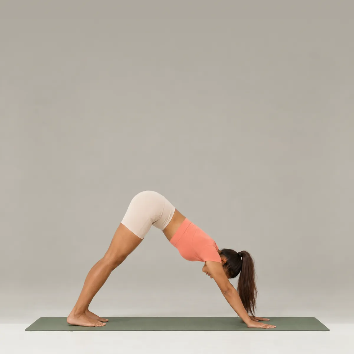 A woman in downward facing dog on a green yoga mat, hips lifted and heels reaching toward the floor