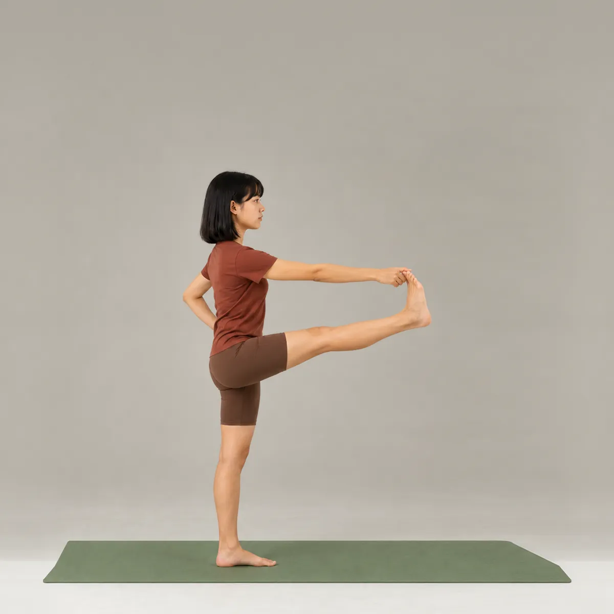 A woman standing on one leg on a green yoga mat, holding her lifted foot out in front of her
