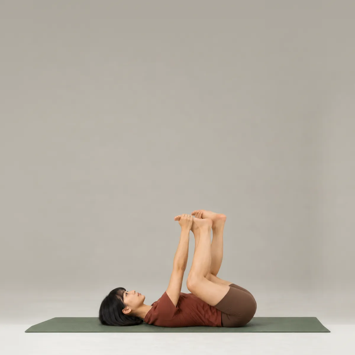 A woman in happy baby pose on a green yoga mat, lying on her back and holding her feet