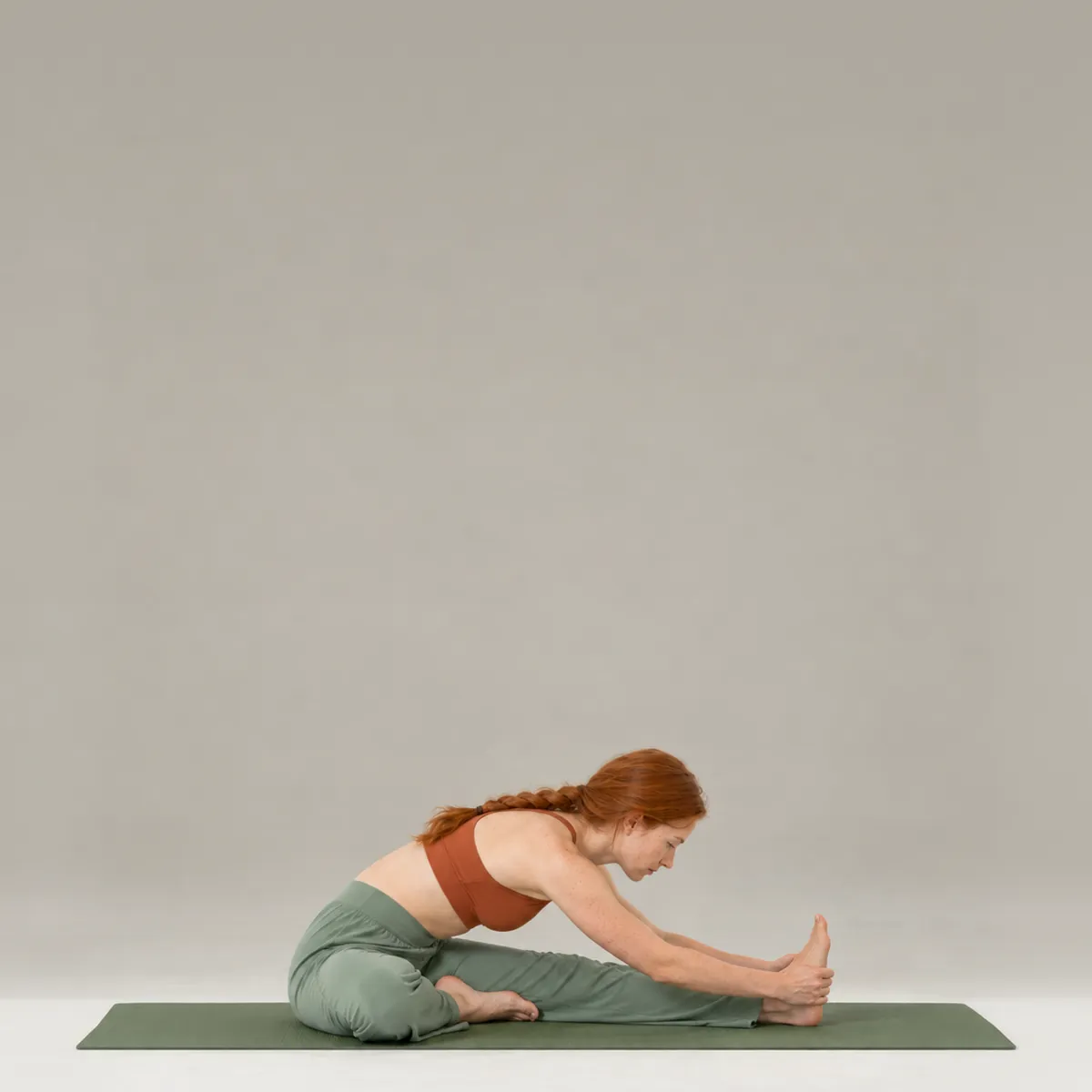 A woman in head to knee pose on a green yoga mat, folding over one extended leg