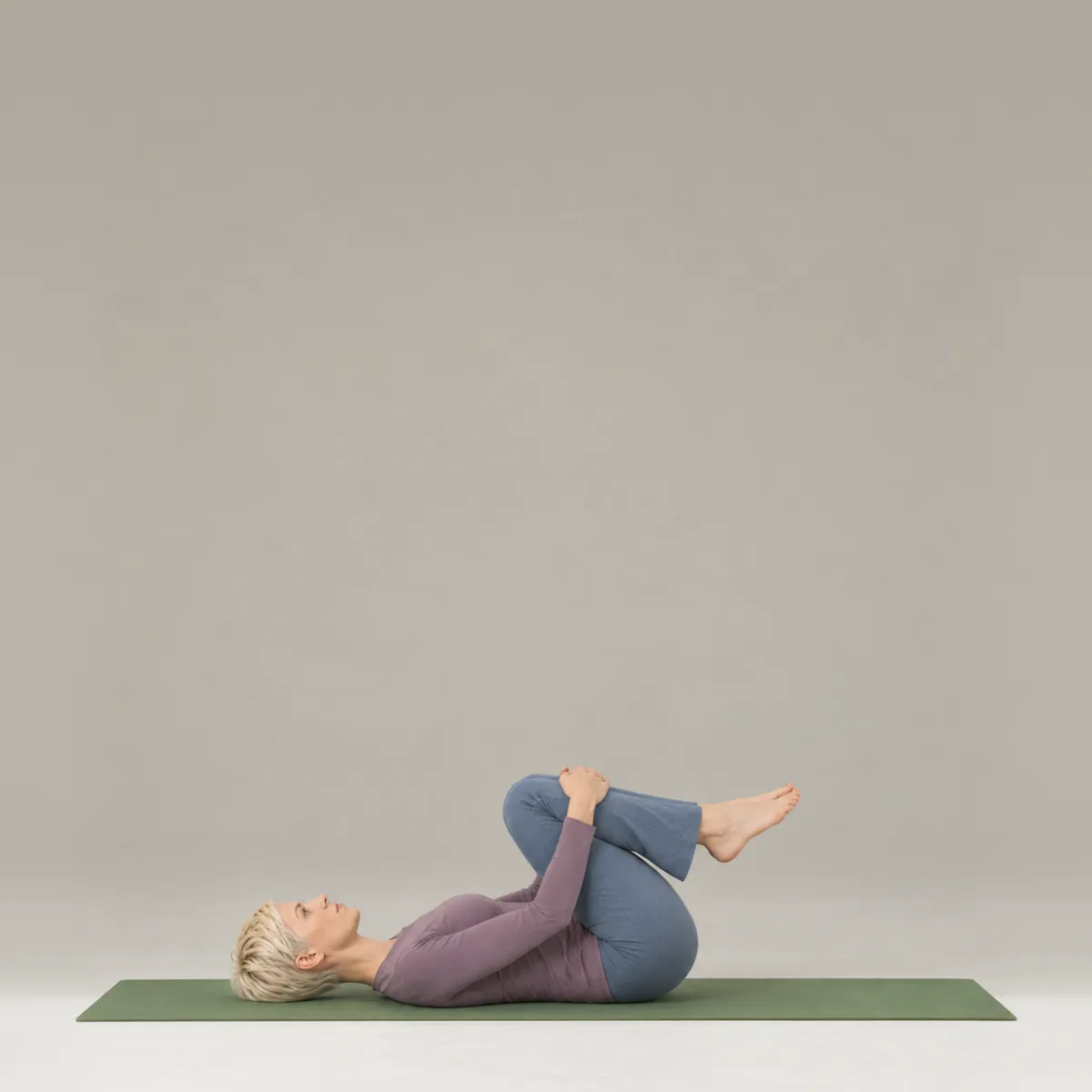 A woman lying on her back on a green yoga mat, hugging both knees toward her chest