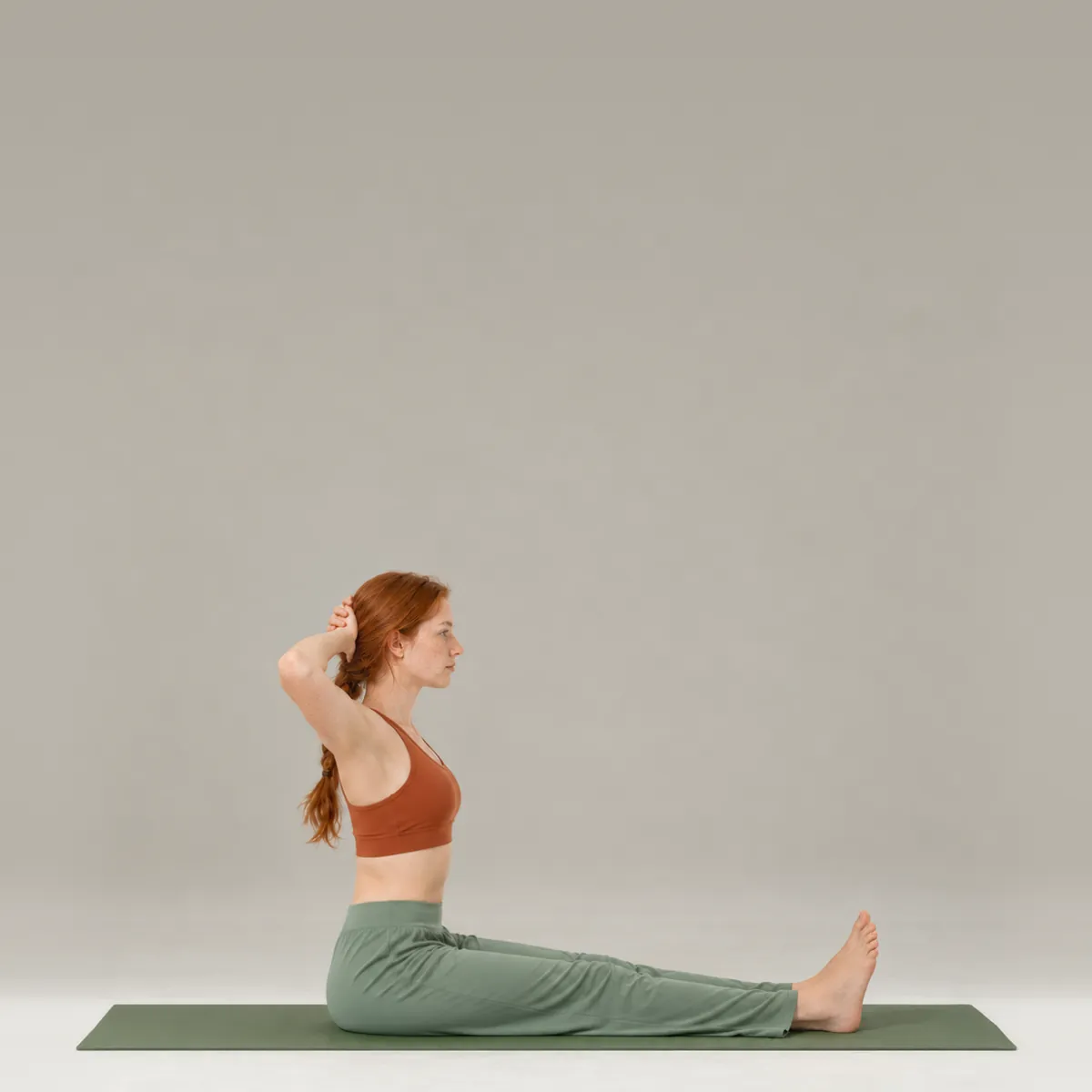 A woman doing the pilates neck pull on a green mat, sitting tall with legs long and hands behind her head