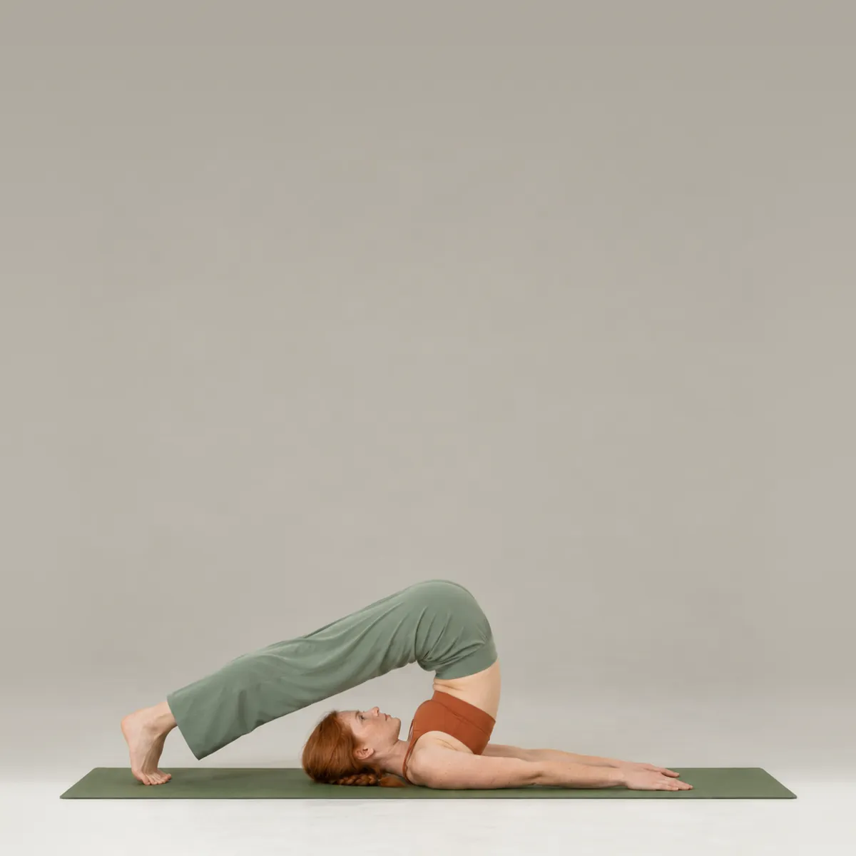 A woman in plow pose on a green yoga mat, legs extended over her head with toes toward the floor