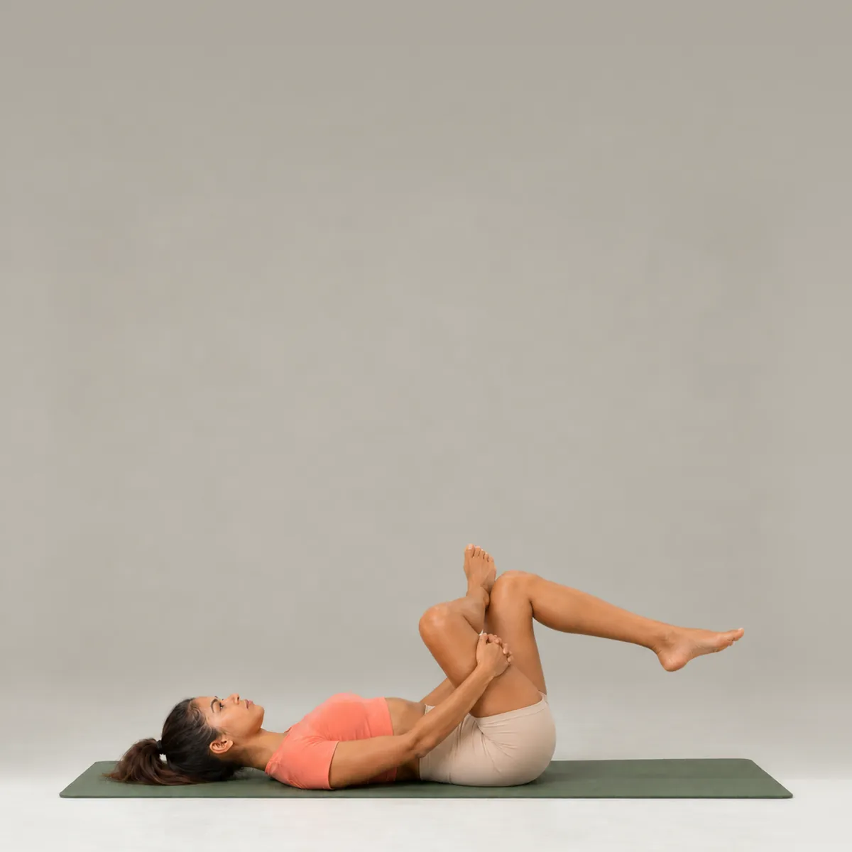 A woman lying on her back on a green yoga mat in a figure four stretch, one ankle crossed over the opposite knee