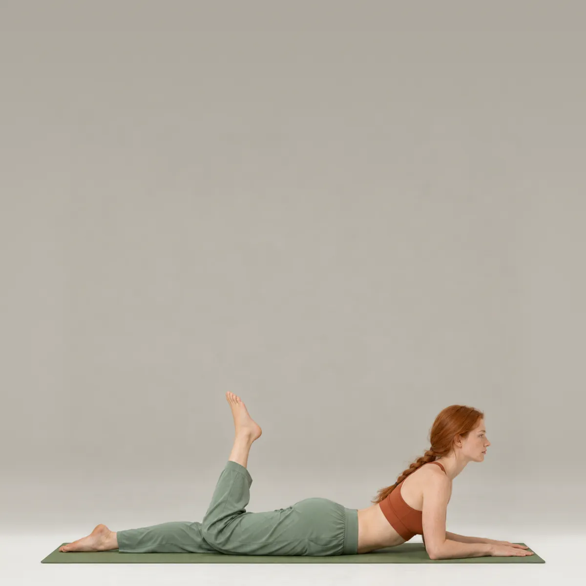 A woman doing the pilates single leg kick on a green mat, lying face down on her forearms with one heel kicking toward her seat