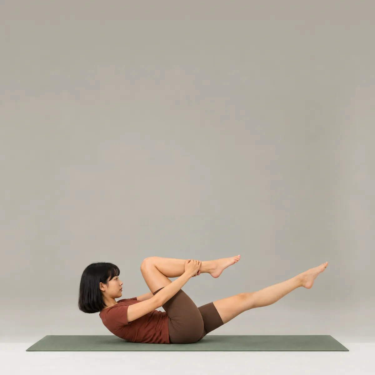 A woman doing the pilates single leg stretch on a green mat, one knee hugged in and the other leg extended