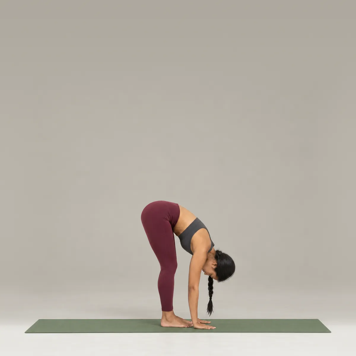 A woman in a standing forward fold on a green yoga mat, folding over her legs with her hands on the mat