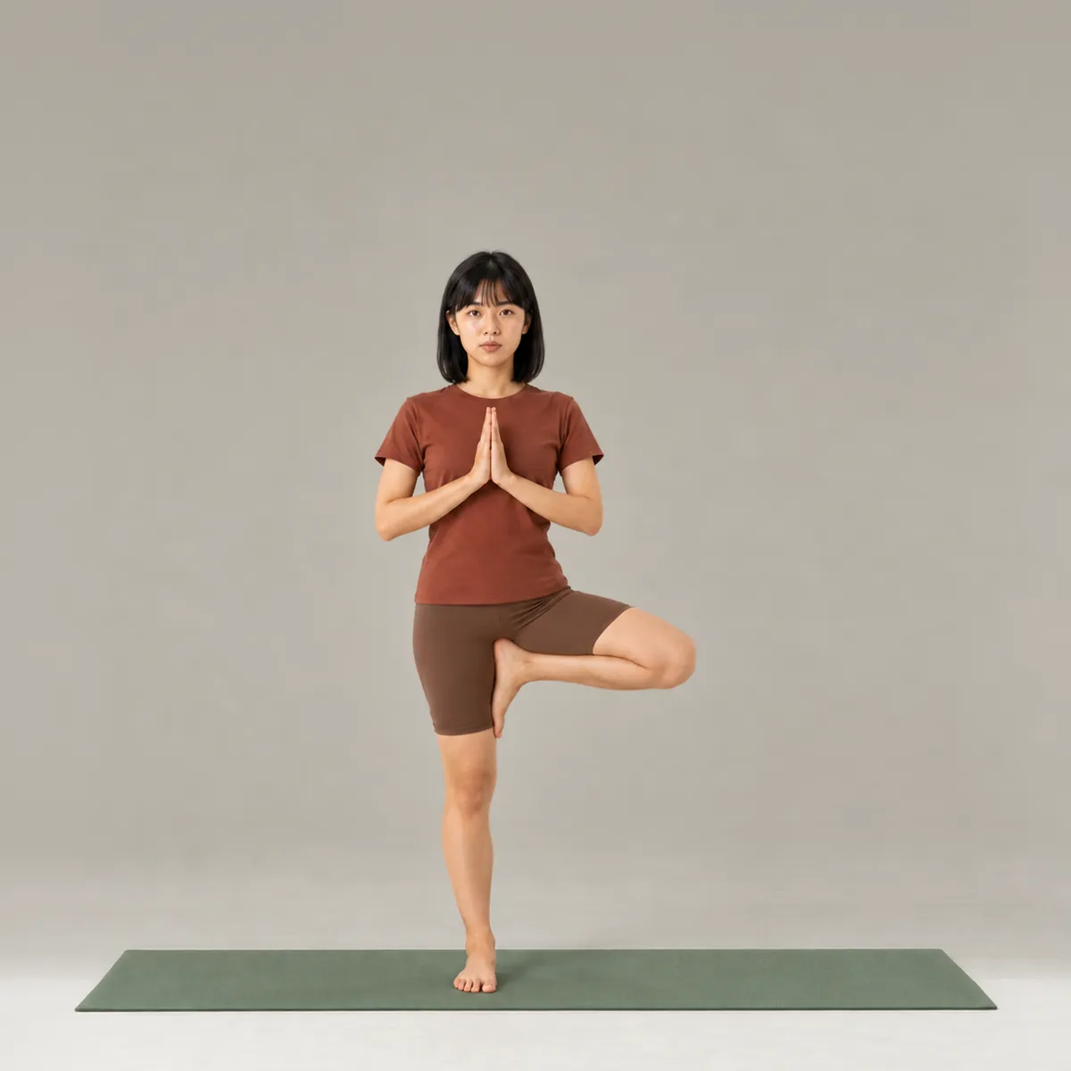 A woman balancing in tree pose on a green yoga mat, one foot on the inner thigh and palms together at her chest