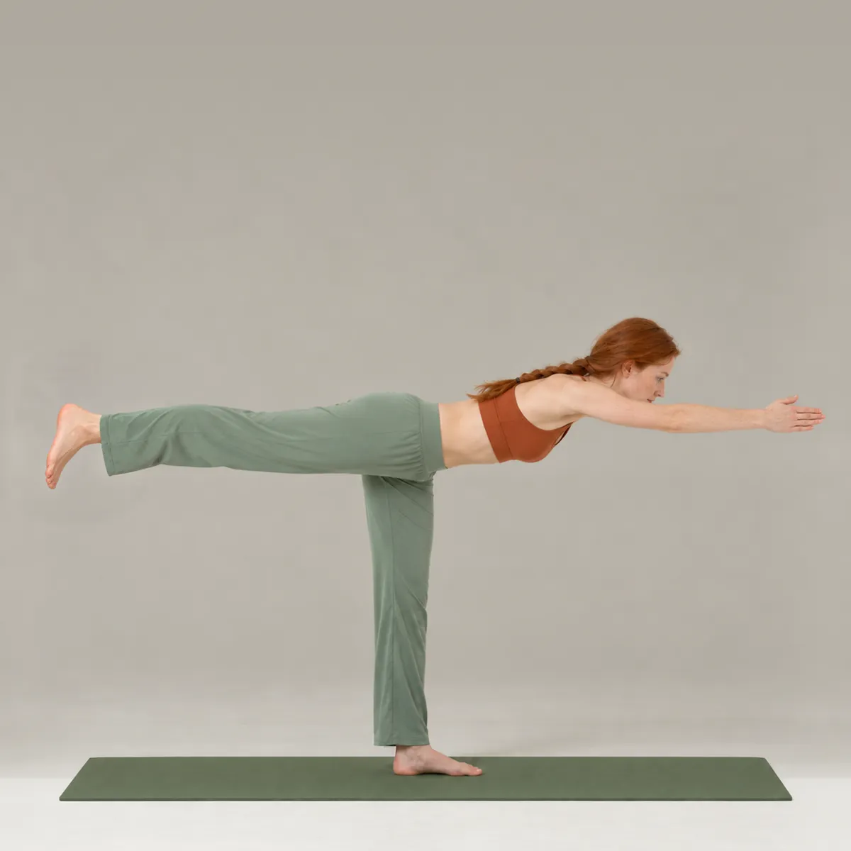 A woman balancing in Warrior III on a green yoga mat, torso and back leg level with the floor